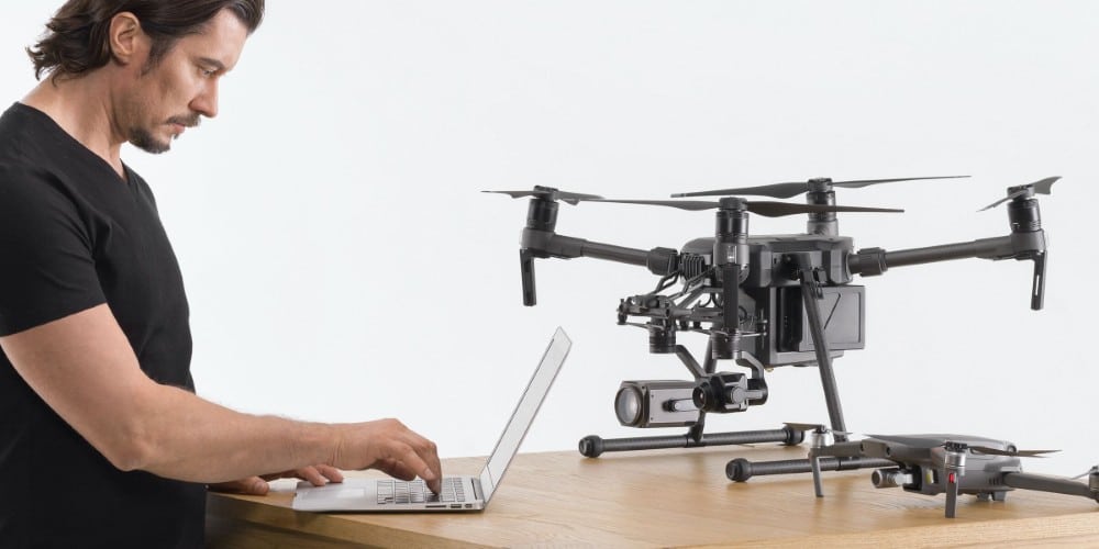 A man in a black T-shirt using a laptop next to a drone on a wooden table.