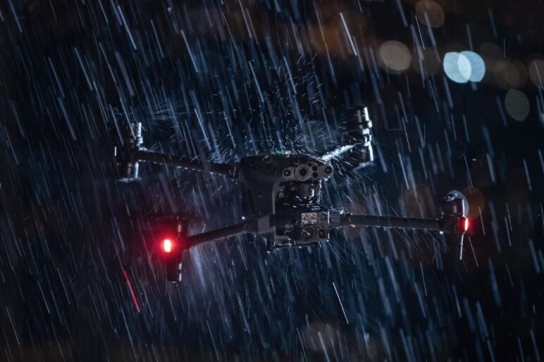 Drone noir avec des lumières rouges en vol sous une pluie intense dans un environnement sombre.