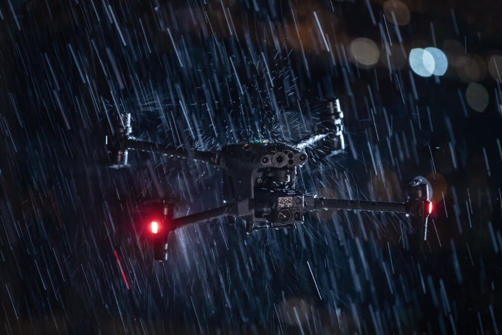 Black drone with red lights flying in intense rain in a dark environment.